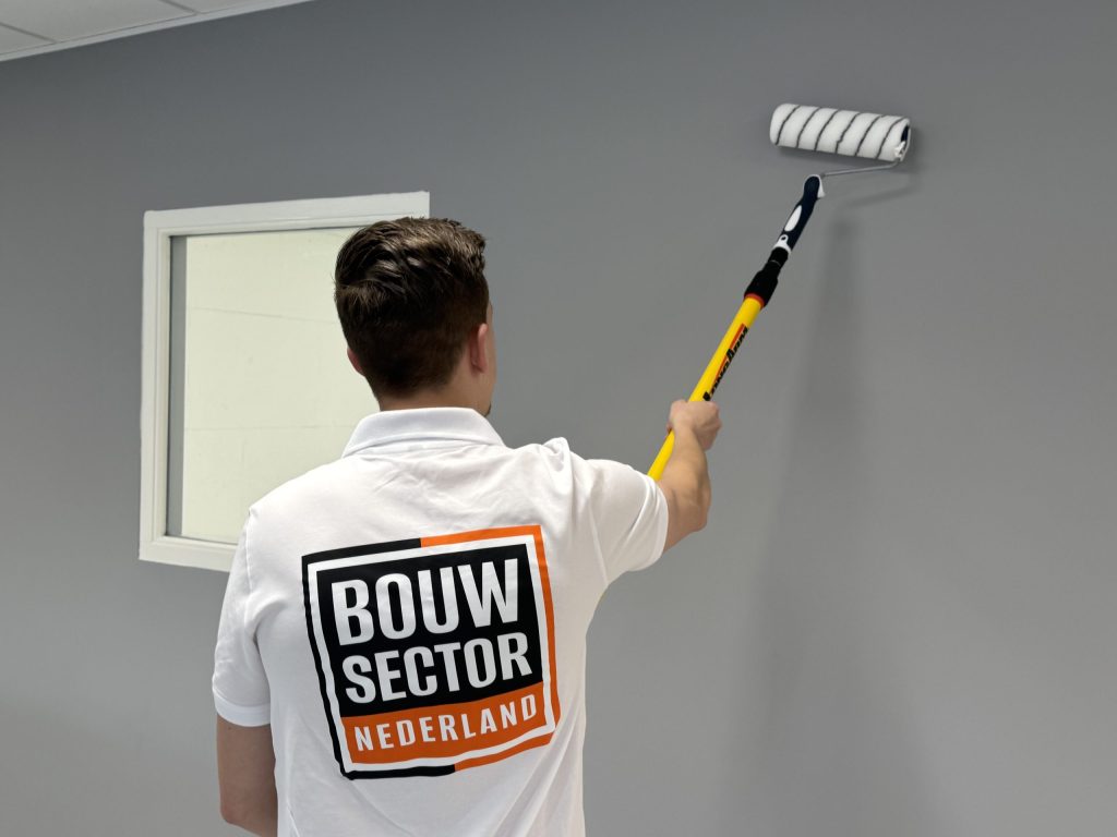 Schilder in wit werkpolo verft een grijze wand vlak naast een wit houten kozijn
