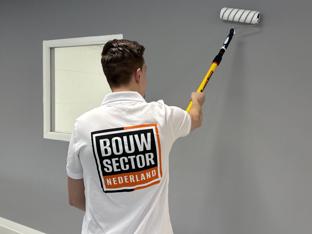 Vakman van Bouwsector Nederland rolt grijze verf op de muur naast een wit raamkozijn