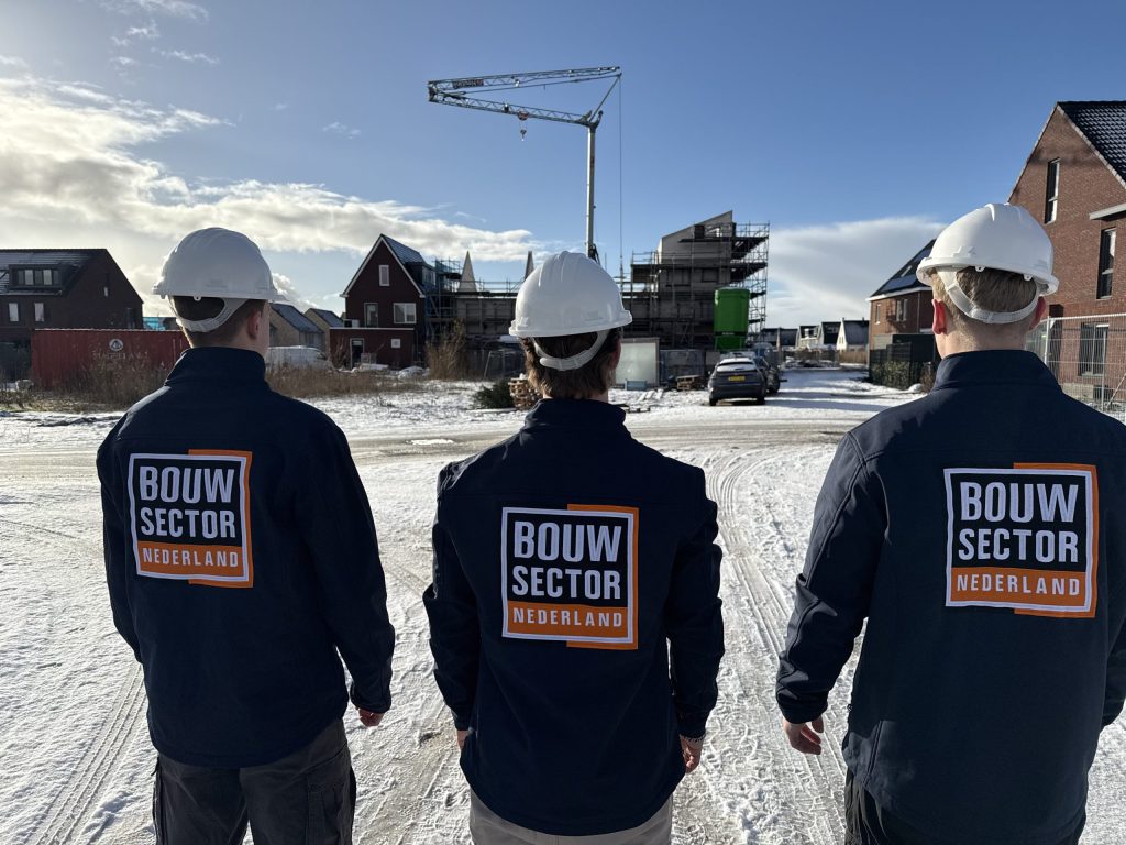 Drie medewerkers in bedrijfsjas met helm op een besneeuwde straat tussen bakstenen woningen