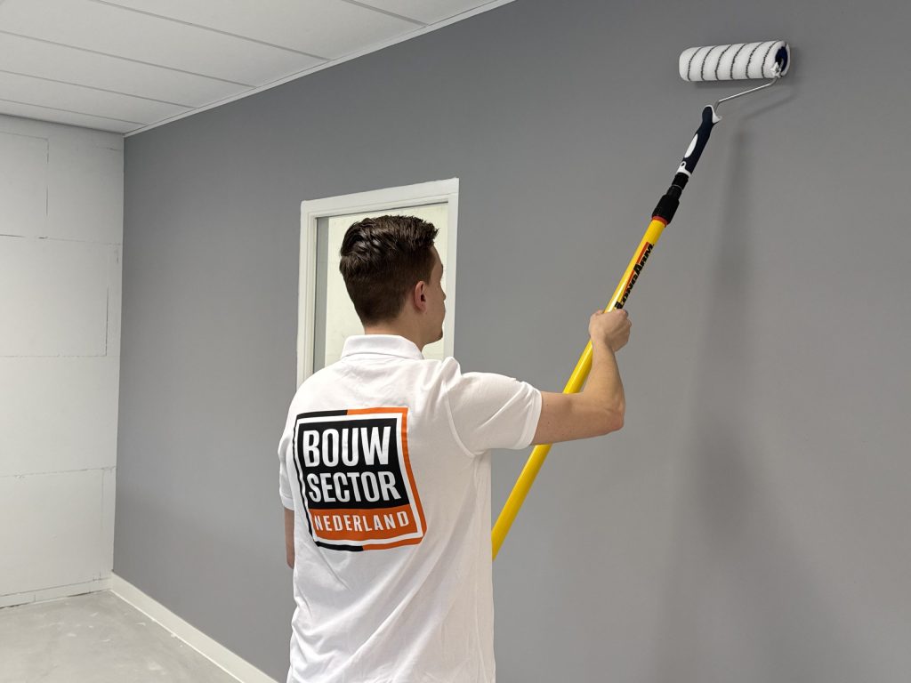 Schilder van Bouwsector Nederland rolt grijze verf op een wand naast een raam, links een kale muur